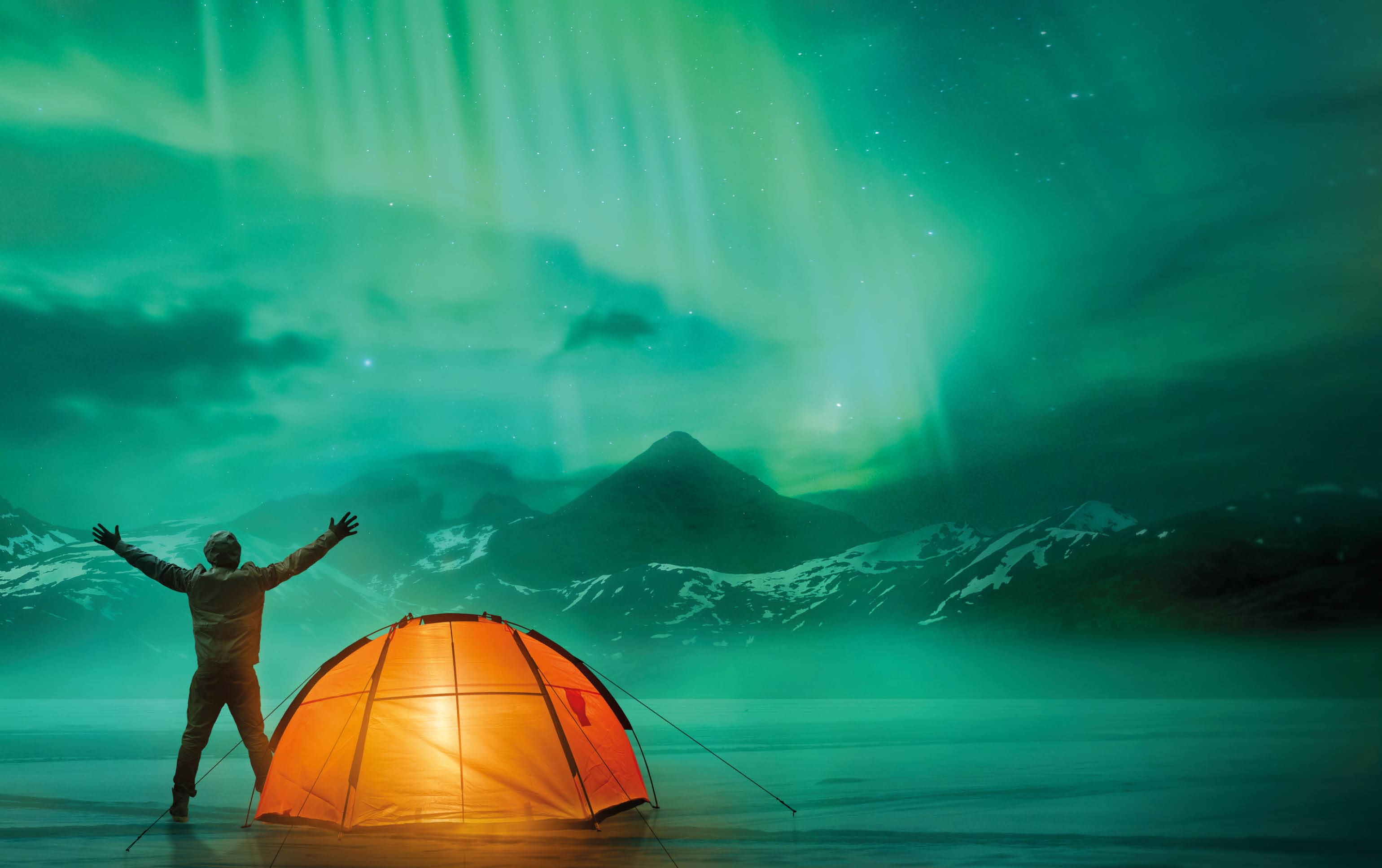 A man camping in wild northern mountains with an illuminated tent viewing a spectacular green northern lights aurora display. Photo composition.
